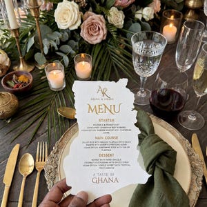 May include: A die-cut menu shaped like the map of Ghana, featuring the text Abena & Kwame Menu, Starter, Main Course, Dessert, and A Taste of Ghana. The menu is held over a place setting with a gold-rimmed plate, a green linen napkin, and gold cutlery.