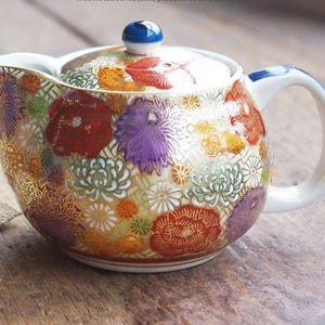 May include: A white ceramic teapot with a floral pattern in shades of red, purple, yellow, and gold. The teapot has a blue knob on the lid and a white handle.