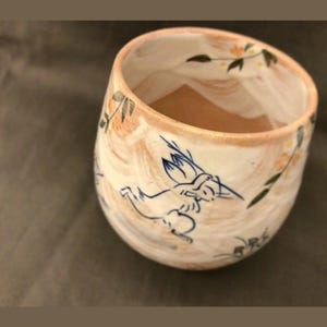 May include: A white ceramic mug with a brown and blue design of a rabbit and flowers. The mug has a rounded shape and a wide rim.