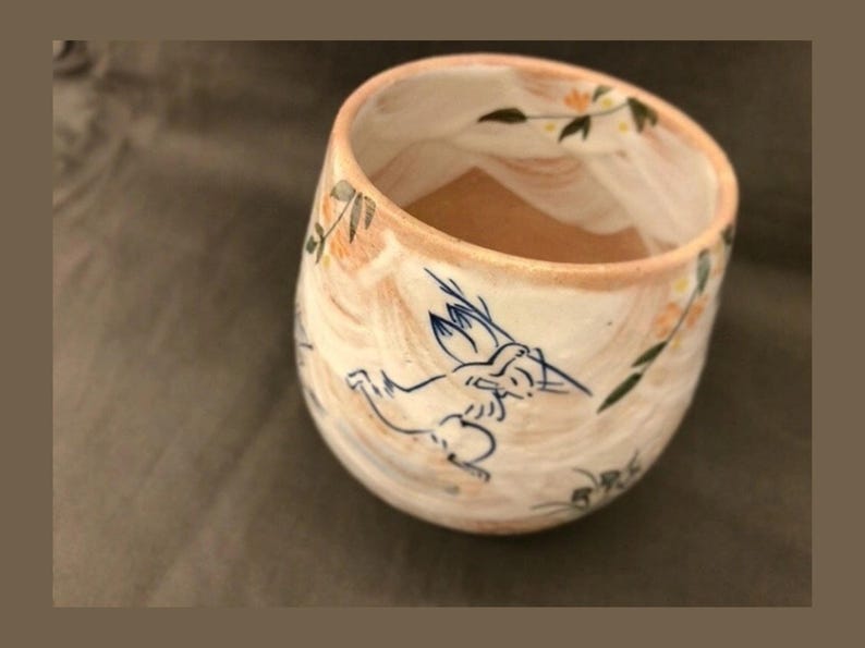 May include: A white ceramic mug with a brown and blue design of a rabbit and flowers. The mug has a rounded shape and a wide rim.