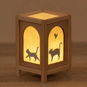 May include: A wooden lantern with a warm, glowing interior. The lantern features arched cutouts with silhouettes of cats and a butterfly. The frame is made of light-coloured wood, and the overall design is simple and elegant.
