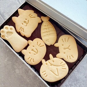 May include: A metal tin filled with six assorted cookies. The cookies are shaped like a cat, a paw print, a fish, and three different cat faces. The cookies are all a light brown colour.