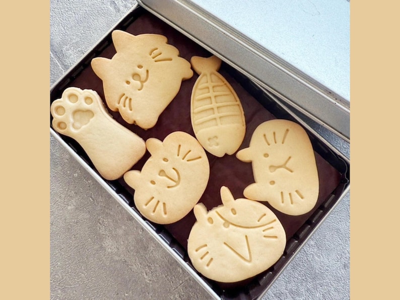 May include: A metal tin filled with six assorted cookies. The cookies are shaped like a cat, a paw print, a fish, and three different cat faces. The cookies are all a light brown colour.