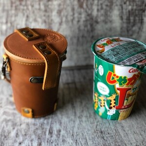 May include: A brown leather cylindrical case with a strap and metal hardware next to a green and white Calbee Jagabee snack cup. The case has a top flap with two straps and a buckle.