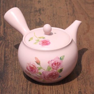 May include: A pink ceramic teapot with a floral design featuring pink roses. The teapot has a spout and a handle.