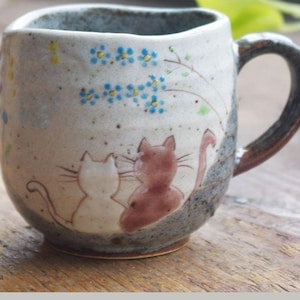 Puede incluir: Una taza de cerámica blanca con un diseño floral azul y amarillo y dos gatos pintados en el lateral. La taza tiene un asa marrón.