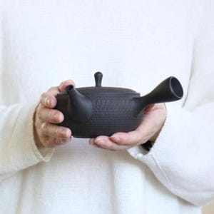 May include: A black ceramic teapot with a textured surface, held in both hands. The teapot has a spout, a handle, and a small lid. The background is a white sweater.
