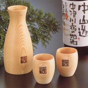 May include: A light-colored wooden sake carafe and two matching wooden cups, each featuring a distinct wood grain pattern and a small square dark brown stamp. The carafe has a narrow neck and a wide base, while the cups are cylindrical and smooth.