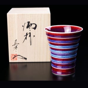 Op de afbeelding: Een rood-blauw gestreepte keramische sake-beker met een houten doos. De doos heeft Japanse karakters erop geschreven.