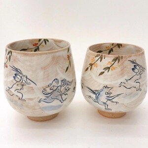 May include: Two ceramic teacups with a light brown glaze and a white and blue floral design. The cups feature a whimsical illustration of rabbits and birds.