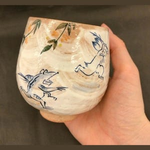 May include: A white ceramic cup with a brown glaze and blue and green painted designs of a frog and a rabbit. The cup is being held in a hand.