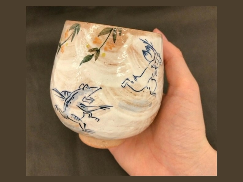 May include: A white ceramic cup with a brown glaze and blue and green painted designs of a frog and a rabbit. The cup is being held in a hand.