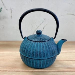 May include: A teal cast iron teapot with a black handle and spout. The teapot has a textured, ribbed design and a matching lid. The teapot is sitting on a wooden surface.