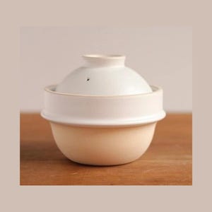 May include: A cream-colored ceramic rice cooker with a matching lid. The lid has a small vent hole. The cooker sits on a wooden surface, with a neutral background. The design is simple and functional.