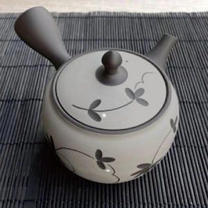Può includere: Una teiera in ceramica con corpo rotondo e manico e beccuccio grigio scuro. Il coperchio è ornato da un pomello grigio scuro e motivi floreali. La teiera è grigio chiaro con accenti floreali grigio scuro.
