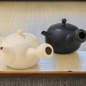 Può includere: Due teiere in ceramica, una color crema e una nera, sono presentate affiancate. La teiera color crema ha una texture maculata, mentre quella nera ha una finitura liscia. Entrambe hanno forma rotonda, un beccuccio e un manico. Le teiere sono su una superficie chiara.