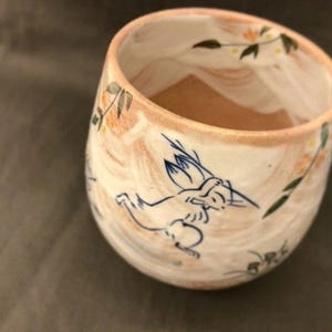 May include: A white ceramic mug with a light brown interior. The mug has a hand-painted design of a stylized animal with blue and brown accents. The design also includes a floral pattern with yellow and green accents.