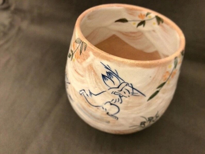 May include: A white ceramic mug with a light brown interior. The mug has a hand-painted design of a stylized animal with blue and brown accents. The design also includes a floral pattern with yellow and green accents.