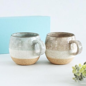 May include: Two ceramic mugs with rounded bodies and attached handles. One mug is light gray, white, and tan, while the other is tan, white, and brown. The mugs are on a white surface with a light blue background.