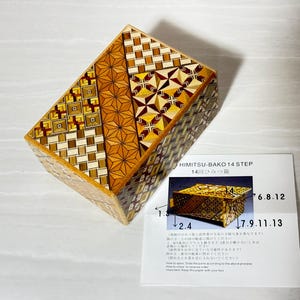 May include: A wooden puzzle box with a geometric pattern in shades of brown and gold. The box is labeled "HIMITSU-BAKO 14 STEP" and "14回ひみつ箱".