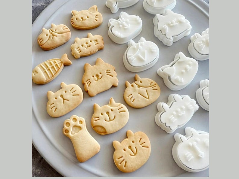 May include: A collection of white plastic cat-shaped cookie cutters and baked cat-shaped cookies on a light grey surface. The cookies are decorated with simple designs, including a fish, a paw print, and a cat face.