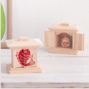 May include: Two wooden miniature shrines. One features a red fish design, the other houses a small figurine. Both are made of light-colored wood and have a traditional Japanese architectural style.