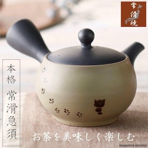 Op de afbeelding: Beige keramische theepot met zwarte handgreep, tuit en deksel. De body heeft een kattenpootafdrukontwerp en een kattenillustratie. Japanse tekst is zichtbaar. Ontworpen om van thee te genieten.