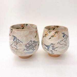 May include: Two ceramic teacups with a white glaze and hand-painted designs. One cup features a floral pattern with a branch of yellow flowers and leaves. The other cup features a whimsical scene of two rabbits running through a field with a bird flying above them.