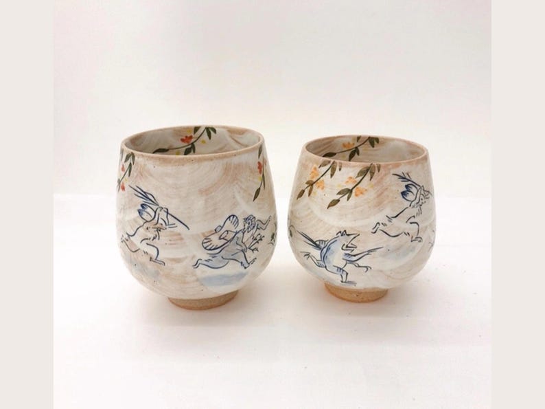 May include: Two ceramic teacups with a white glaze and hand-painted designs. One cup features a floral pattern with a branch of yellow flowers and leaves. The other cup features a whimsical scene of two rabbits running through a field with a bird flying above them.