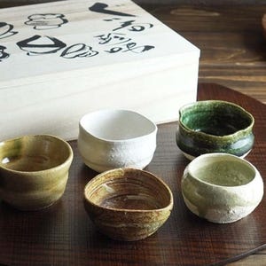 May include: Set of six small ceramic bowls in various colours and glazes, including green, brown, white, and a speckled brown glaze. The bowls are arranged on a wooden surface.