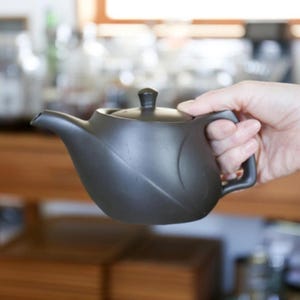 Op de afbeelding: Een donkergrijze keramische theepot met een gebogen lichaam, tuit en handvat. De theepot heeft een klein, rond deksel en wordt in de hand van een persoon gehouden. De achtergrond is wazig, wat wijst op een keuken of eetkamer.