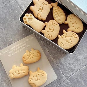 May include: A tin box filled with various cat-shaped biscuits. The biscuits are light brown and have different designs, including a paw print, a fishbone, and a cat face. The box is silver and has a white label with the text "A gift full of love and spiced with care. Here is quality time. We are with gentle time."