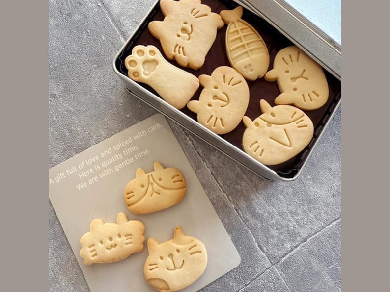 May include: A tin box filled with various cat-shaped biscuits. The biscuits are light brown and have different designs, including a paw print, a fishbone, and a cat face. The box is silver and has a white label with the text "A gift full of love and spiced with care. Here is quality time. We are with gentle time."
