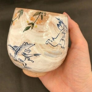 May include: A white ceramic teacup with a brown glaze and blue and green painted designs of birds and a rabbit. The cup is being held in a hand.
