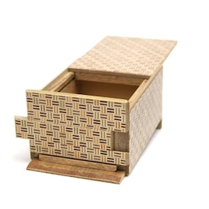 Puede incluir: Una caja de rompecabezas de madera con un cajón deslizante y una tapa con bisagras. La caja está hecha de madera clara con un patrón geométrico.