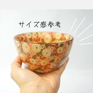 Puede incluir: Un cuenco melocotón con motivos florales crema y verdes es sostenido en una mano. El diseño del cuenco presenta flores y hojas estilizadas. El texto japonés "サイズ感参考" es visible.