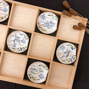 Può includere: Un vassoio di legno con una griglia di scomparti contenente sei piccoli piatti rotondi in ceramica. Ogni piatto presenta un'illustrazione dipinta a mano di una rana o di un uccello con accenti floreali blu, verdi e arancioni. Il vassoio è posto su uno sfondo scuro.