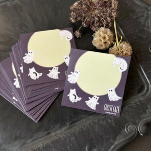 Może przedstawiać: Stos kwadratowych fioletowych karteczek samoprzylepnych z jasnożółtym okręgiem i białymi ilustracjami kotów-duchów. Słowa "GHOST CATS" są wydrukowane w prawym dolnym rogu notatki. Suche kwiaty i torebki nasienne w tle.