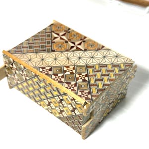 Puede incluir: Una caja de madera con un intrincado patrón geométrico hecho de maderas de diferentes colores. La caja está cerrada y tiene un pequeño pestillo de madera en el lateral.