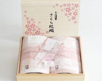 Puede incluir: Dos toallas rosas con un patrón blanco están dobladas y atadas con una cinta rosa en una caja de madera. La tapa de la caja tiene un diseño floral y el texto "さくら タオル 2枚組" en japonés.