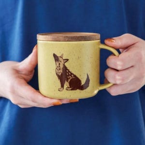 Puede incluir: Una taza de cerámica amarilla con una tapa de madera presenta una ilustración marrón y blanca de un lobo aullando. La taza tiene un asa y está siendo sostenida por la mano de una persona.