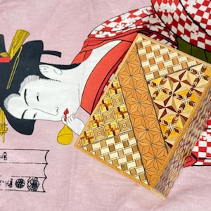 Puede incluir: Una caja de rompecabezas de madera con intrincados patrones geométricos en tonos marrones y dorados. La caja está abierta y el interior es visible. La caja está sentada sobre una tela rosa con un diseño de estilo japonés.