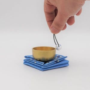 Puede incluir: Un pequeño cuenco tibetano dorado con un percutor esférico transparente suspendido sobre él. El cuenco descansa sobre una pila de cuadrados de tela azul con un patrón geométrico blanco y azul. La imagen está sobre un fondo blanco.