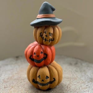 May include: A stacked Halloween decoration featuring three carved pumpkins. The top pumpkin wears a black witch hat with an orange band. The pumpkins are orange and tan with black facial features. The decoration is a festive autumn or Halloween item.
