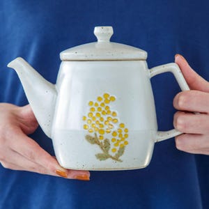 Könnte beinhalten: Eine weiße Teekanne aus Keramik mit einem Blumendesign mit gelben Blüten und grünen Blättern. Die Teekanne hat einen Deckel und einen Griff.