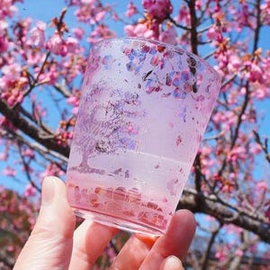 May include: A clear, pink-tinted glass with a floral design. The glass features an etched tree and flower pattern, with pink and purple accents. The background shows a blurred view of pink cherry blossoms and a blue sky.