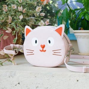 Puede incluir: Un bolso bandolera rosa claro con forma de gato con detalles de orejas naranjas, ojos negros y nariz roja. El bolso tiene una correa larga y ajustable y una cremallera plateada. El bolso está sobre una superficie blanca con plantas al fondo.