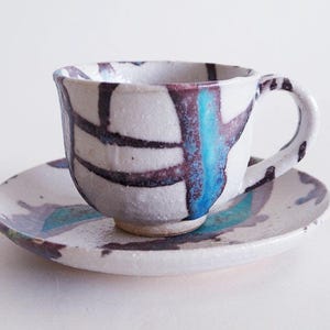 Könnte beinhalten: Eine weiße Teetasse und Untertasse aus Keramik mit abstrakten blauen und violetten Pinselstrichen.