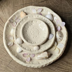 Può includere: Un set di tre piatti in ceramica bianco sporco di diverse dimensioni, adornati con conchiglie e fiori rosa secchi. I piatti sono disposti in una configurazione nidificata, creando un'esposizione decorativa. I piatti sono su una superficie di legno.