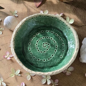 Puede incluir: Un cuenco de cerámica hecho a mano con un interior verde vibrante y un borde blanco roto texturizado. El cuenco presenta un diseño en espiral en el centro y está rodeado de elementos decorativos, incluyendo flores y conchas. El cuenco tiene unos 15 cm de diámetro.